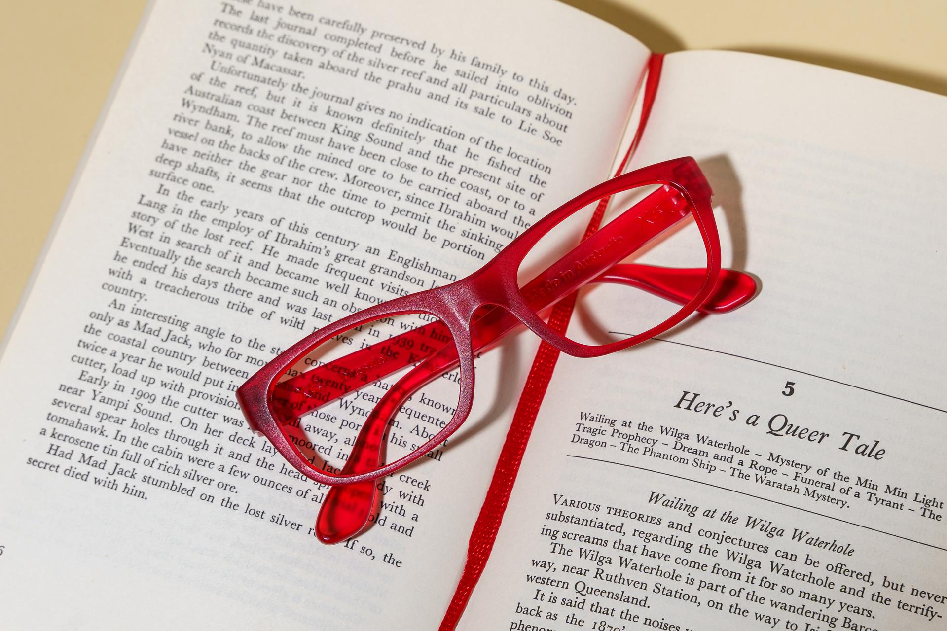 Red reading glasses rest on an open book, hinting at the relief they offer from headaches caused by reading.