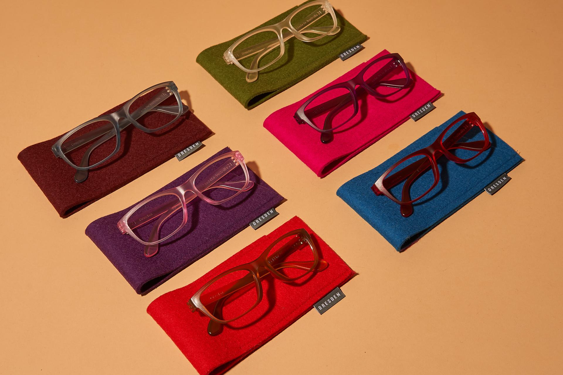 A colorful flat lay of six pairs of Dresden Vision eyeglasses, each resting on a vibrant, matching felt pouch. The glasses feature various translucent and matte frames in shades of grey, pink, orange, green, purple, and red, arranged diagonally on a neutral tan background under soft, warm lighting.