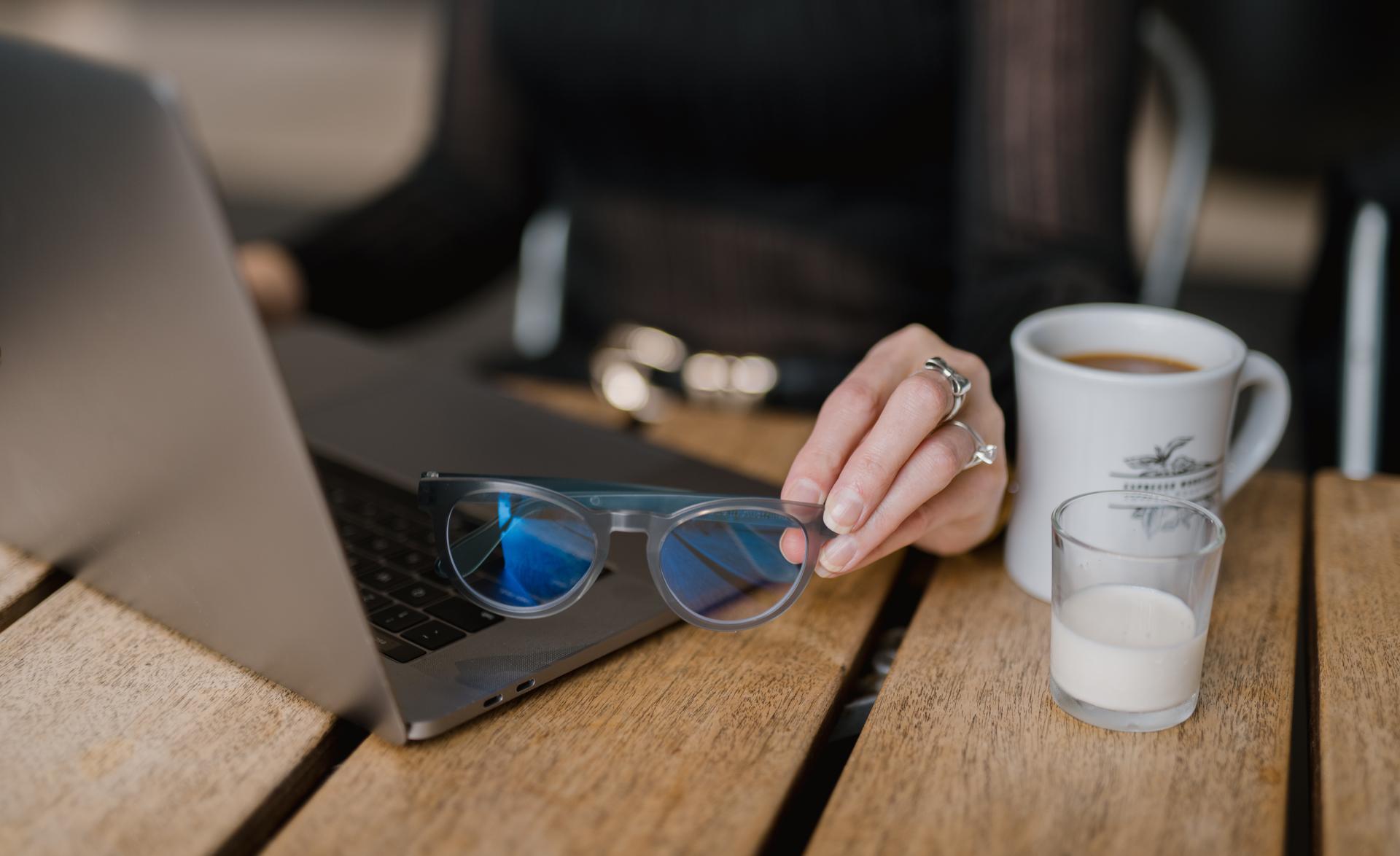 A person multitasks with coffee and a laptop while holding prescription glasses, reflecting modern eyewear needs at Dresden Vision Canada.