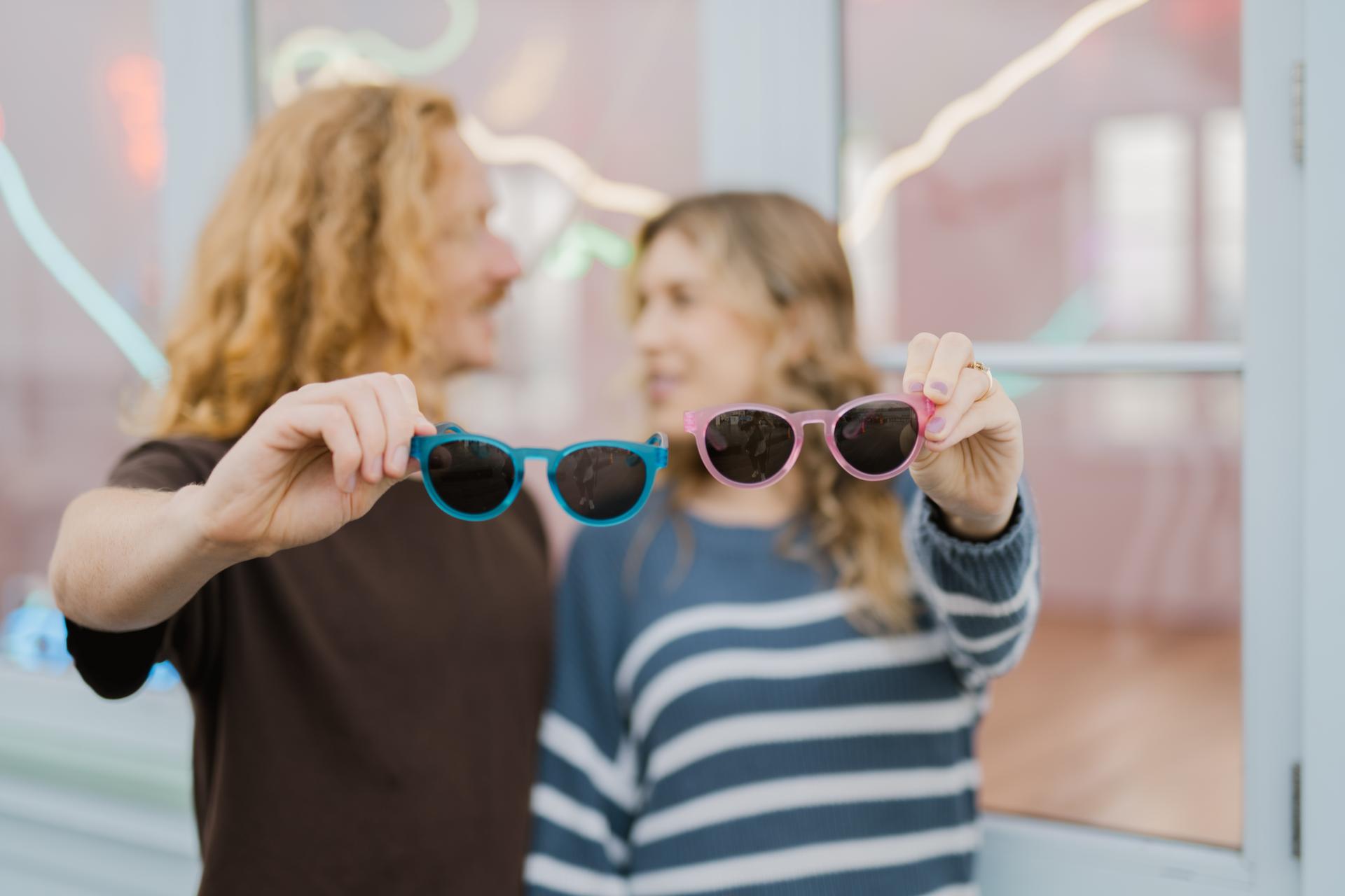 A couple holding up colourful sustainable sunglasses while smiling at each other, used in Dresden Vision Australias guide to types of multifocal lenses for sunglasses that improve clarity and comfort outdoors.