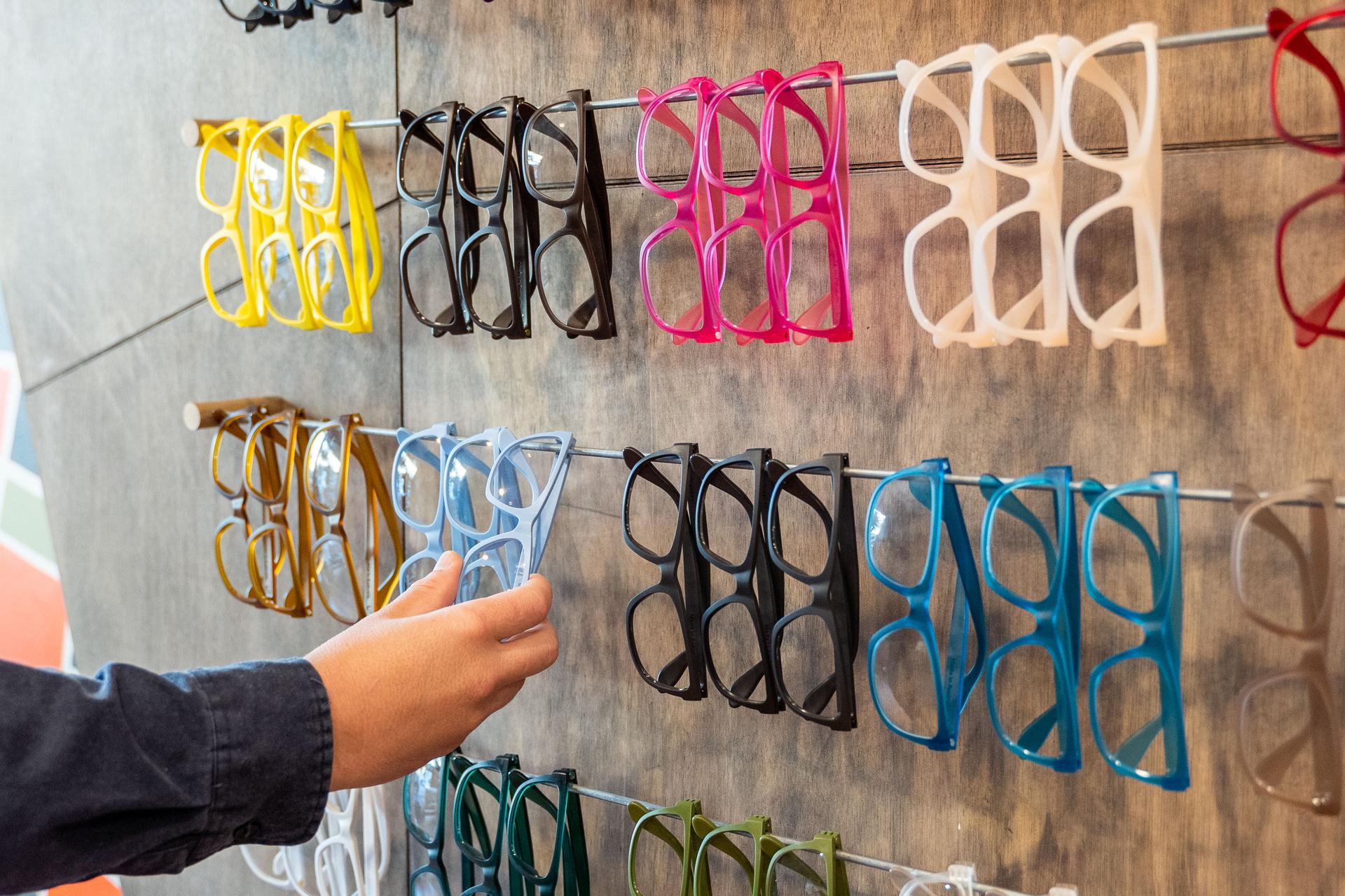 A person’s hand reaching for a pair of light blue frames from a vibrant wall display featuring Dresden Vision Canada eyeglasses in an array of colors, including yellow, pink, black, and teal.