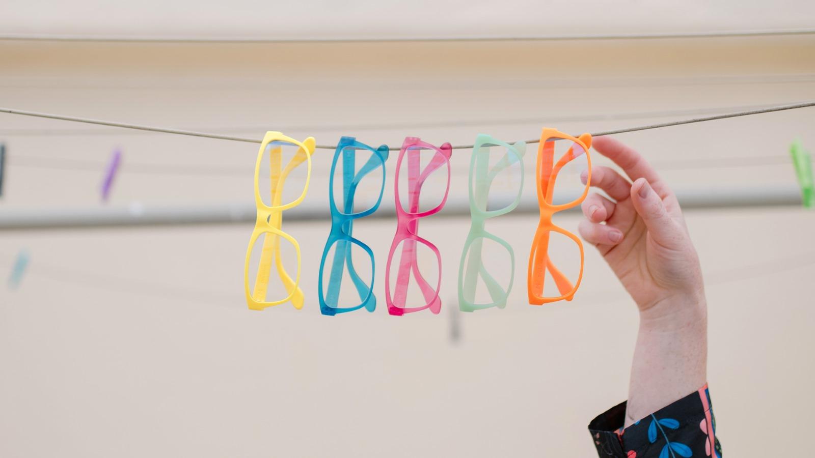 A hand reaches for one of five vibrant Dresden Vision eyeglasses (yellow, blue, pink, green, orange) hanging on a clothesline. This image showcases the playful and customizable eyewear available from Dresden Vision Australia.