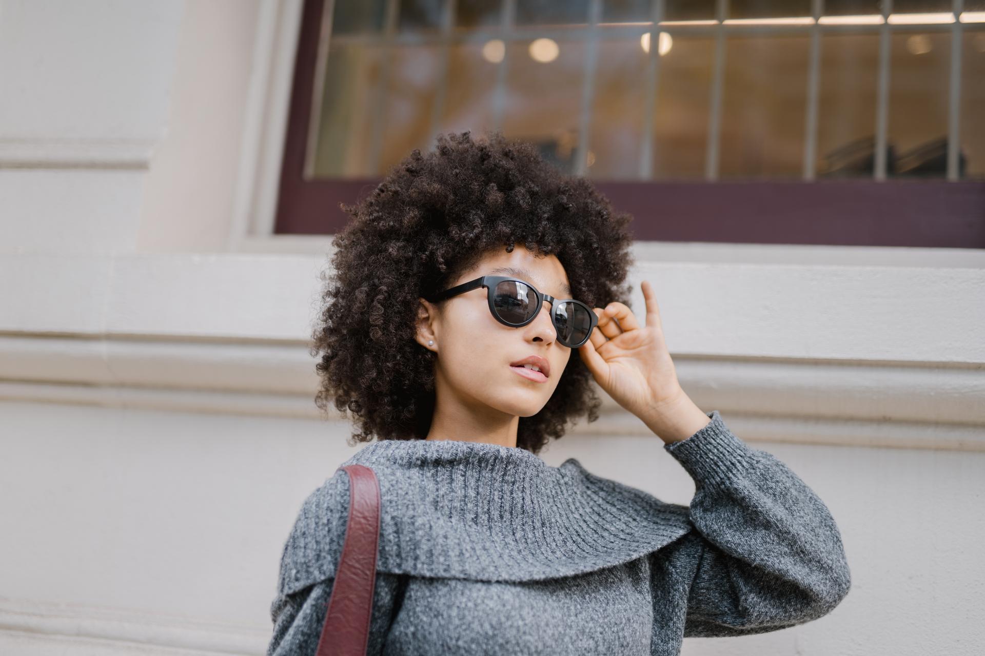 Close-up of a woman with dark, voluminous curly hair, seen from the side, with her hands near her temples adjusting her dark sunglasses. This image is courtesy of Dresden Vision Canada.