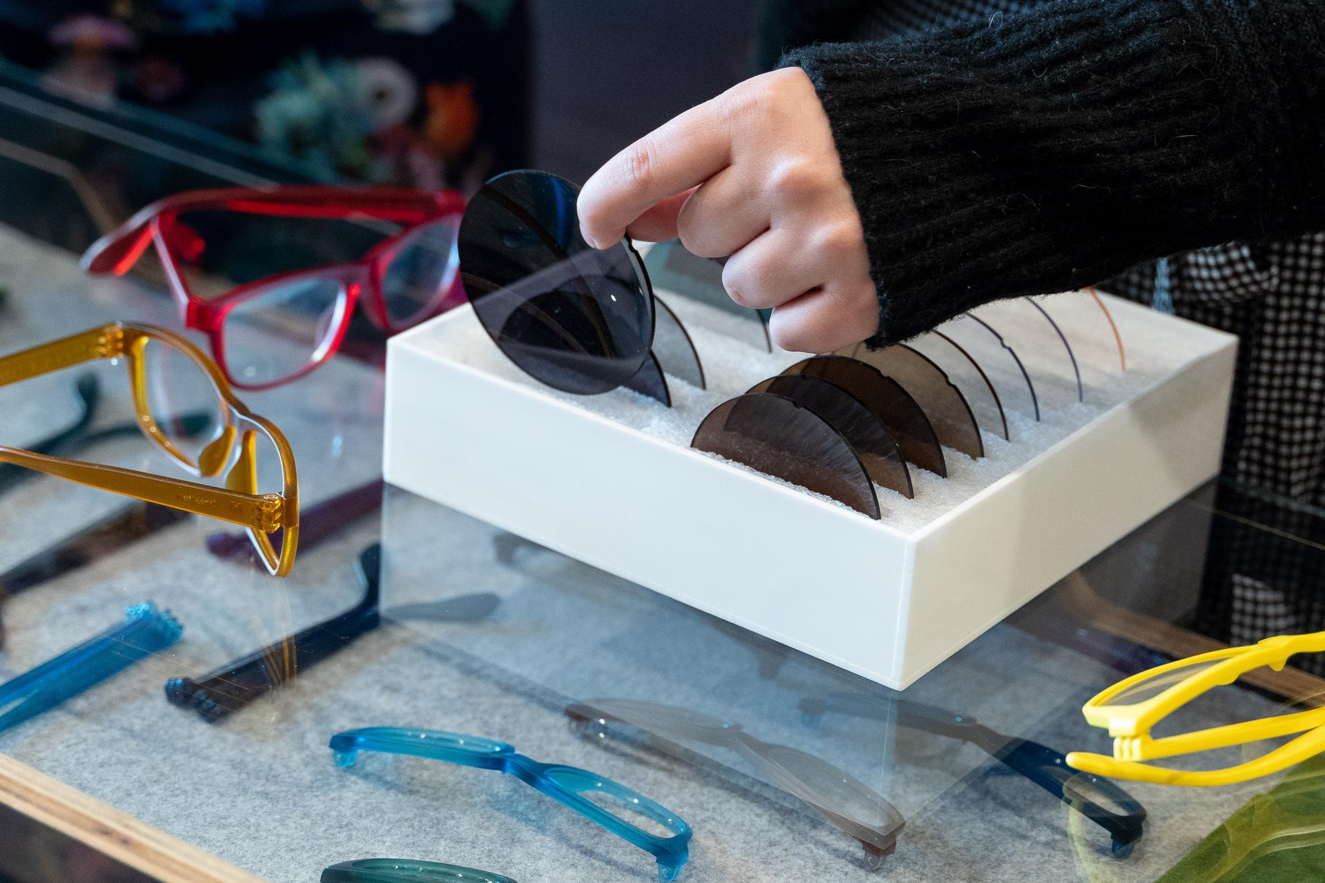 A hand choosing a sunglass lens from a sample set at Dresden Vision United States, with eco-friendly frame options scattered on the glass surface.