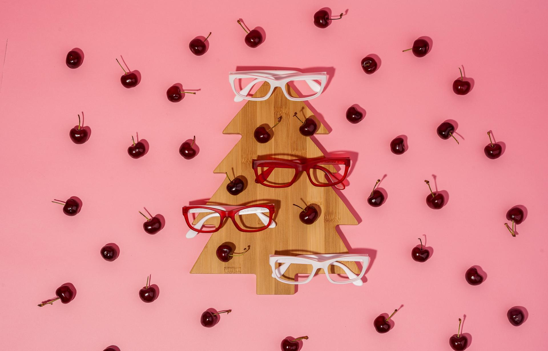 Festive flat lay of red and white Dresden Vision Australia eyeglass frames arranged on a wooden Christmas tree board with cherries on a pink background, celebrating Boxing Day with style and sustainability.