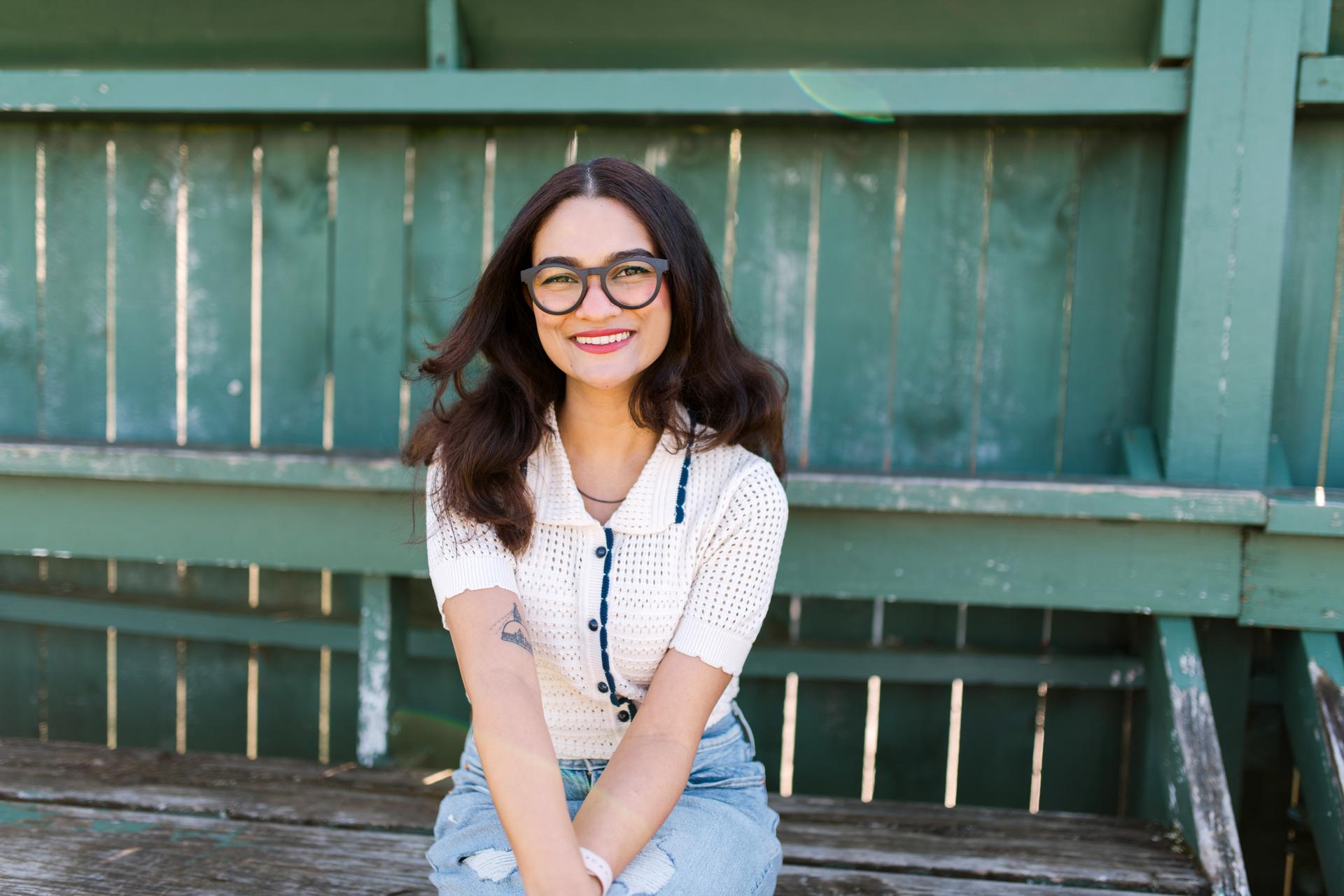 Young woman smiling in round Dresden Vision New Zealand glasses, seated in front of a green bench, representing clear vision and sustainable eyewear ideal for student study sessions.