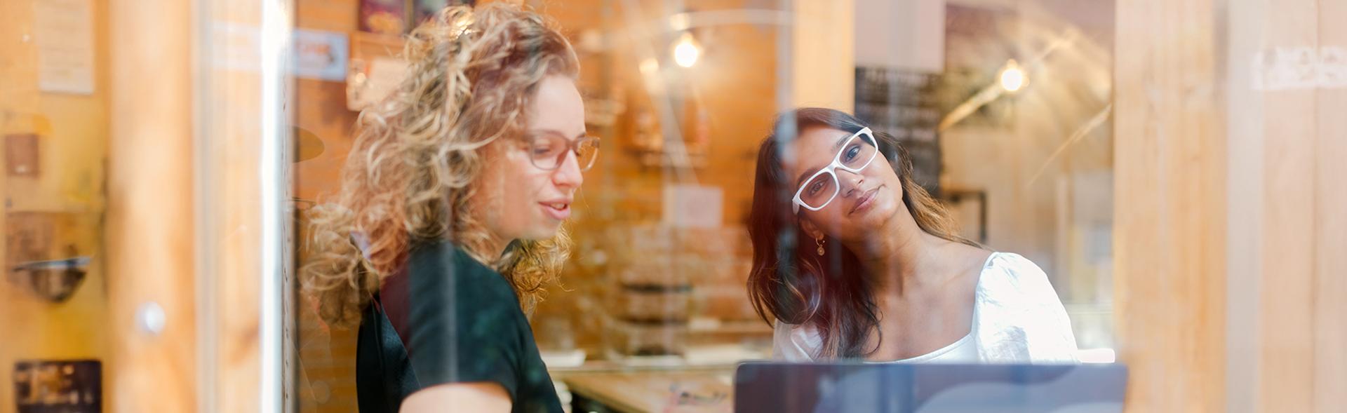 Two women wearing stylish Dresden Vision Australia glasses collaborate at a café, viewed through a window with reflections of a cozy interior.