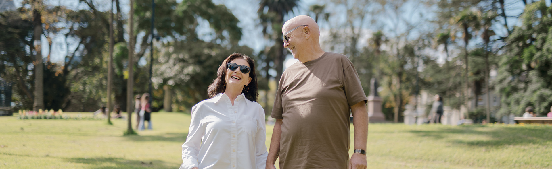 A smiling elderly couple wearing stylish sunglasses holds hands while walking in a sunny park, captured in a joyful and candid moment – Dresden Vision Australia.