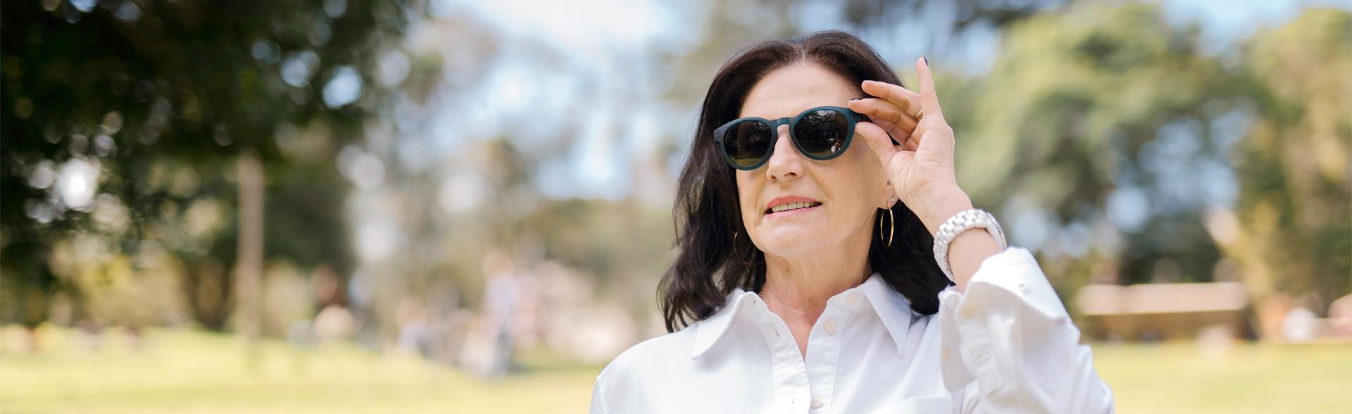 Confident older woman wearing Polarised Sunglasses in Australia, dressed in a white shirt, smiling outdoors in a sunny park, enjoying clear vision and stylish eye protection under bright daylight.
