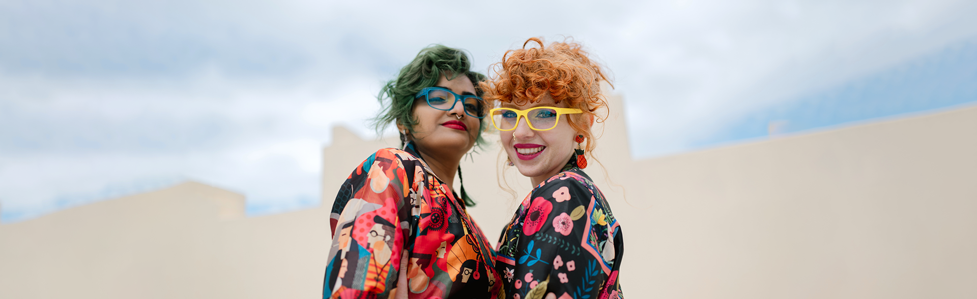 Wearing vibrant Dresden Vision New Zealand womens glasses, two friends stand close, dressed in colorful patterns and sharing a happy moment.