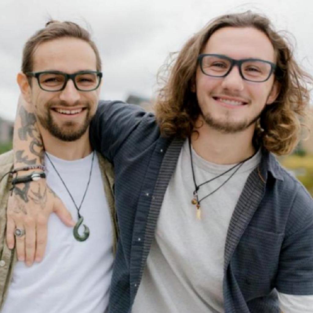 Two men wearing affordable men's glasses in Australia, standing side by side in an outdoor urban area with a background of street art, greenery, and distant buildings, both smiling and dressed casually.