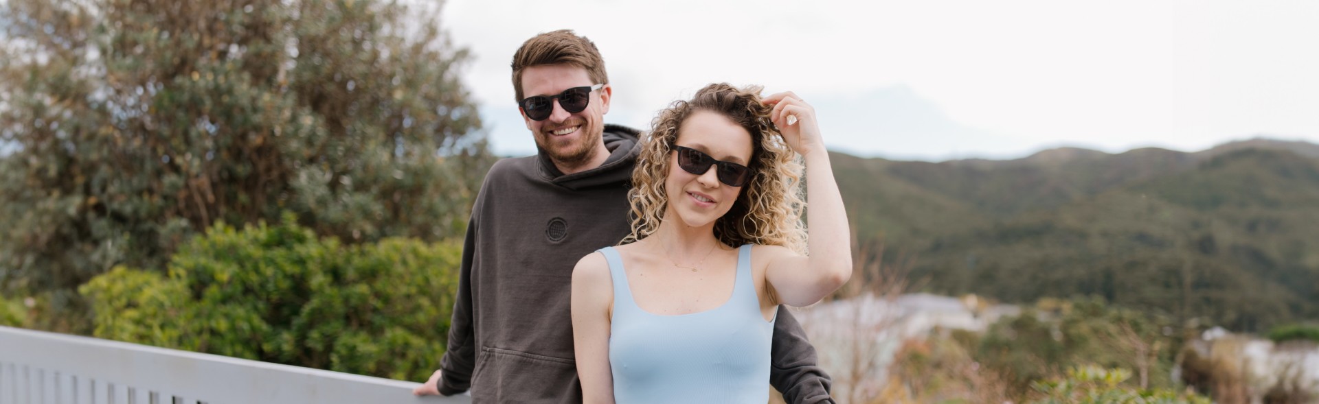 A stylish couple wearing Dresden Vision International sunglasses, enjoying the beauty of nature with rolling green hills and trees in the background. The woman, dressed in a light blue sleeveless top, adjusts her sunglasses while the man in a dark hoodie leans back on a white railing, exuding effortless style.