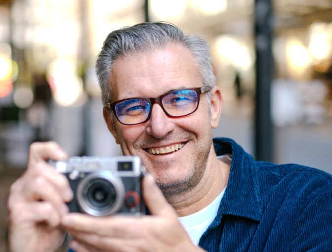 Man wearing Dresden Vision blue light glasses, smiling while holding a camera in a warmly lit indoor setting