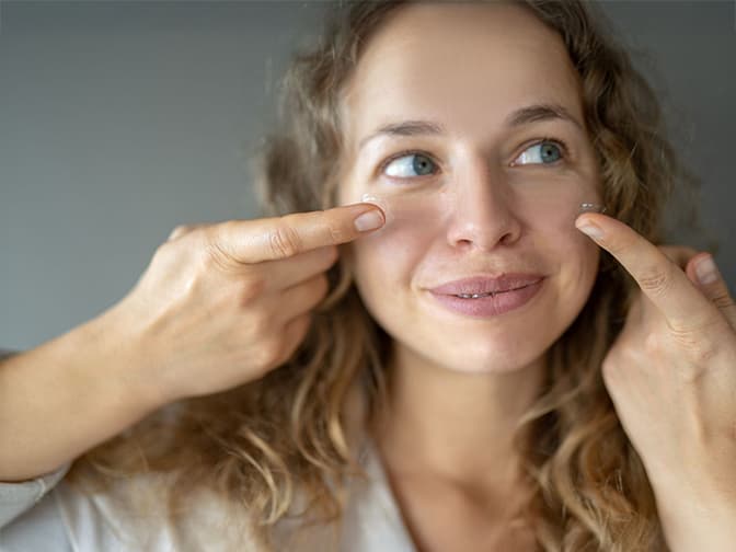 A smiling woman with curly hair holding up a pair of contact lenses on her fingers, highlighting the contact lens collection at Dresden Vision Canada.