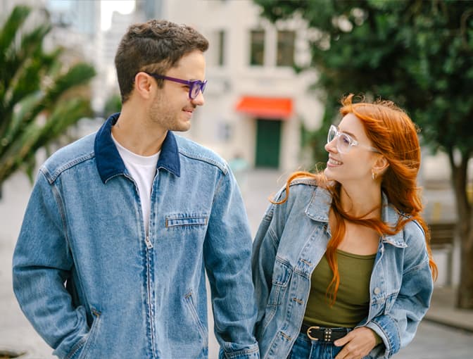 Young couple wearing Dresden Vision prescription glasses, smiling at each other outdoors, man in violet frames and woman in clear frames