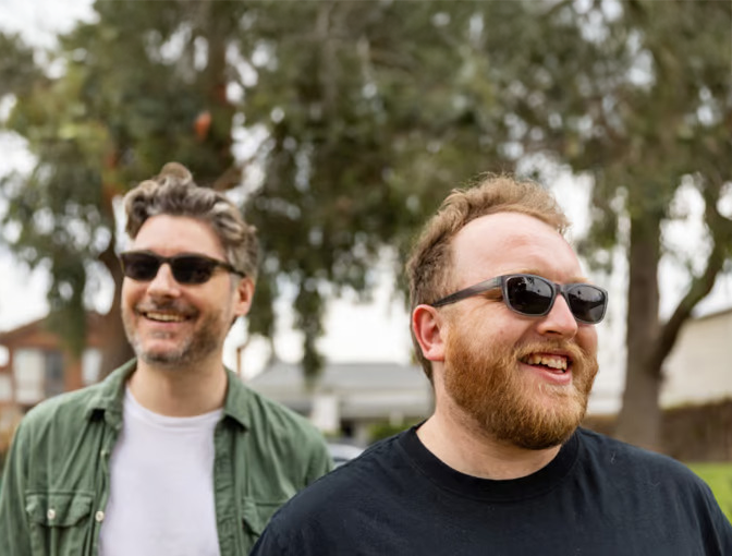 Two men wearing prescription sunglasses in Australia, enjoying a sunny day outdoors. The man on the left has grey hair, a beard, and is dressed in a green polo with a white shirt, while the man on the right has a ginger beard and is sporting a black T-shirt.