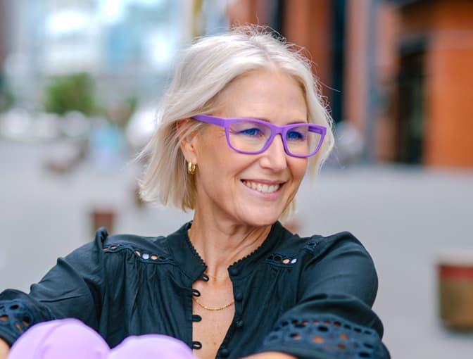 Woman wearing purple Dresden Vision progressive glasses, smiling outdoors in a bright urban environment