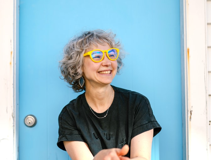 Woman in Dresden Vision Canada reading glasses, wearing bold yellow frames and looking off to the side against a blue backdrop