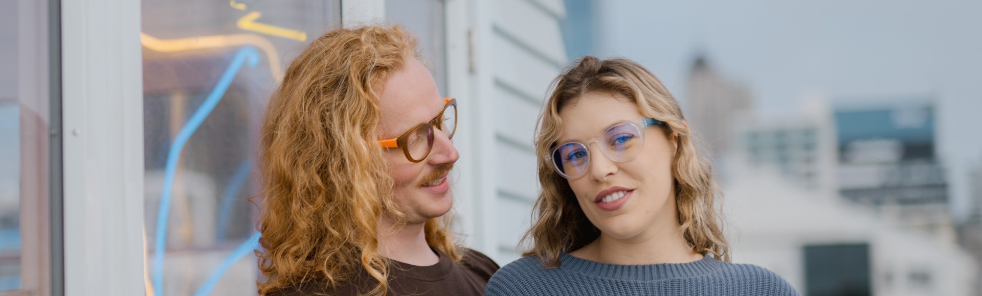 Friends stand close, modeling clear and orange Dresden Vision Canada prescription glasses with relaxed smiles and a soft city skyline behind them.