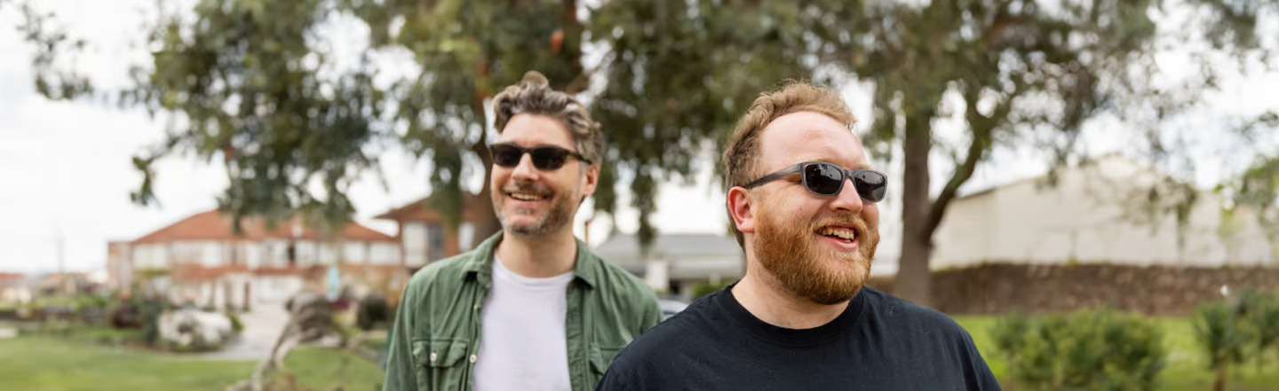 Two men enjoying a sunny day outdoors in the United States, both wearing prescription sunglasses. The man on the left has grey hair and a beard, in a green polo and white shirt, while the man on the right has a ginger beard and a black T-shirt.