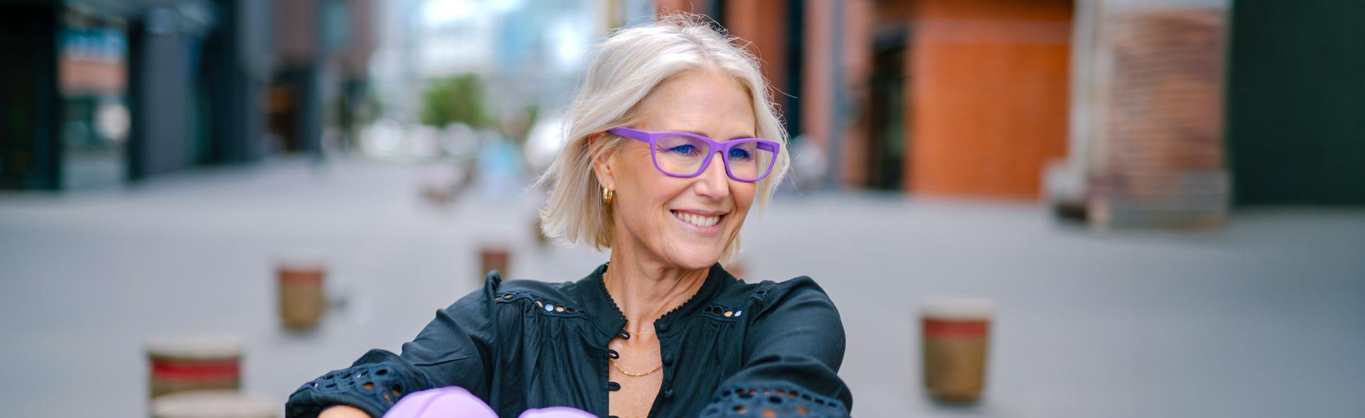 Woman wearing purple Dresden Vision progressive glasses, smiling outdoors in a modern city setting