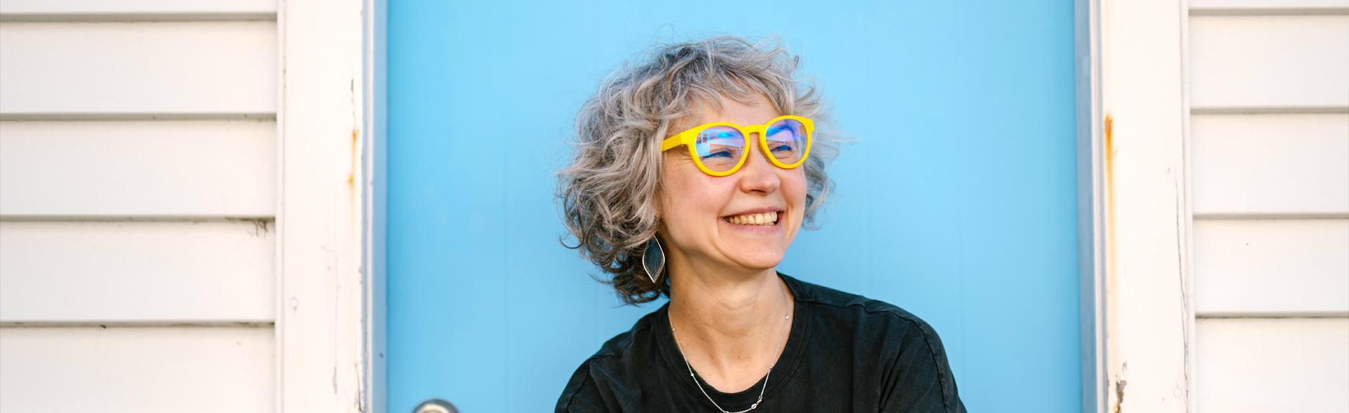 Smiling woman wearing Dresden Vision New Zealand reading glasses, featuring yellow frames in a vibrant outdoor setting