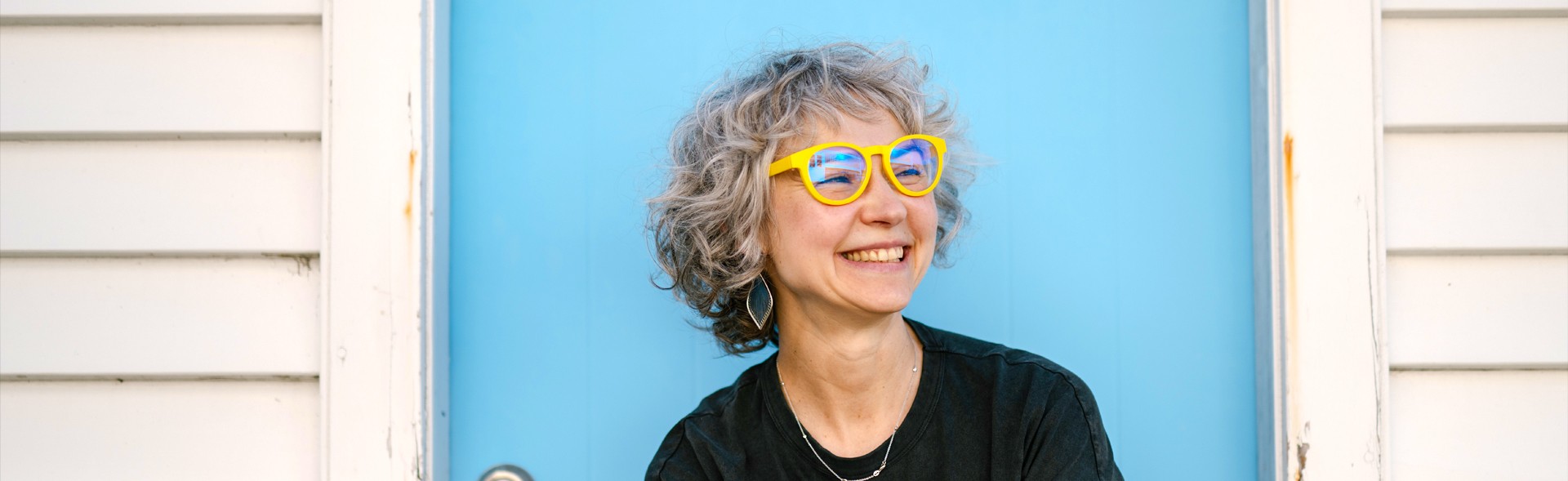 Woman in Dresden Vision Canada reading glasses, wearing bold yellow frames and looking off to the side against a blue backdrop