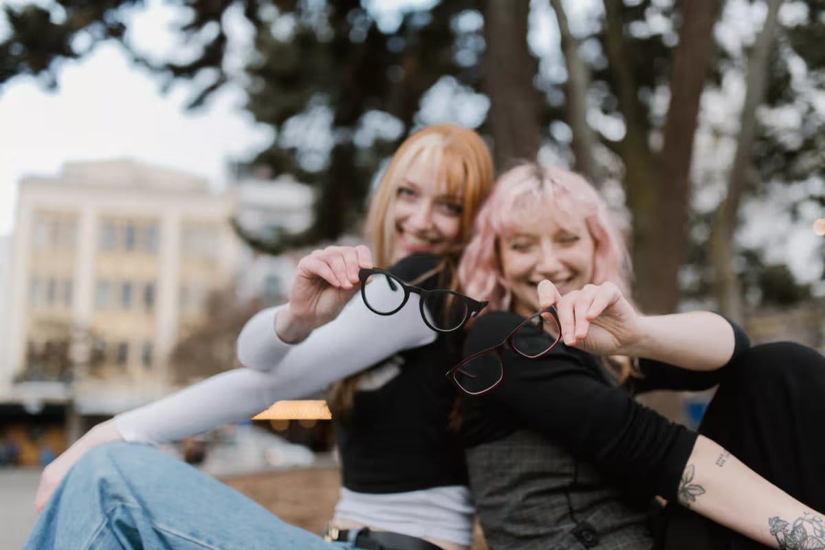 Smiling duo showing off their colourful frames with Single Vision Lenses from Dresden Vision New Zealand, enjoying a laid-back day beneath city trees.