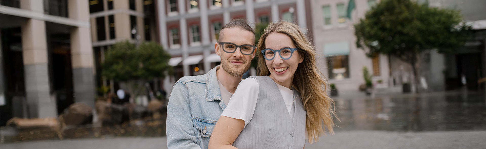 A smiling couple on a city street, both wearing matching blue rectangular and round shaped eyeglasses. He is in a light denim jacket, while she wears a white outfit and her blonde hair flows in the wind, featuring Dresden Vision United States.