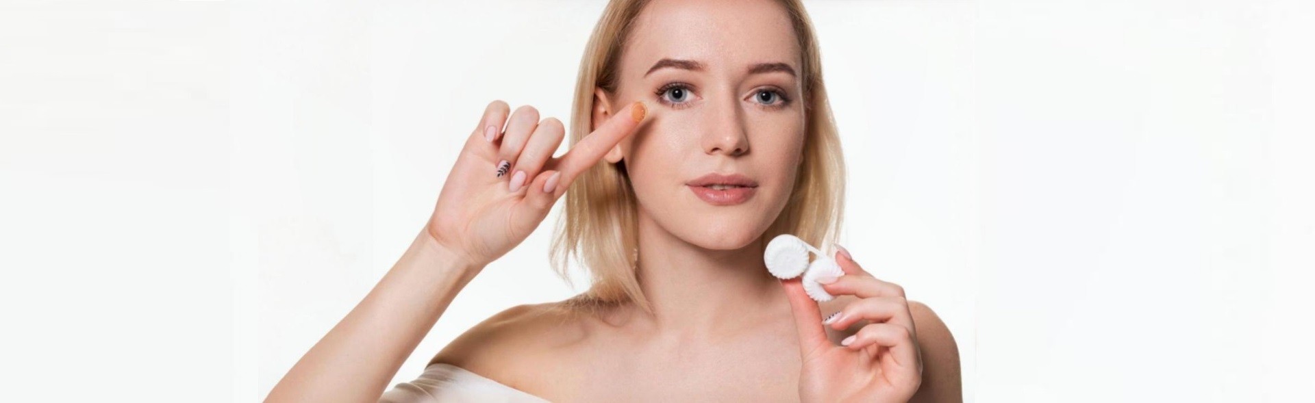 Young woman with blonde hair inserting a contact lens. She is holding her eyelid open with one hand while bringing a contact lens towards her eye with the other. In her other hand, she holds a contact lens case. The woman is wearing a white top and has a focused expression against a plain light background. This image showcases Dresden Vision Canada contact lens products.