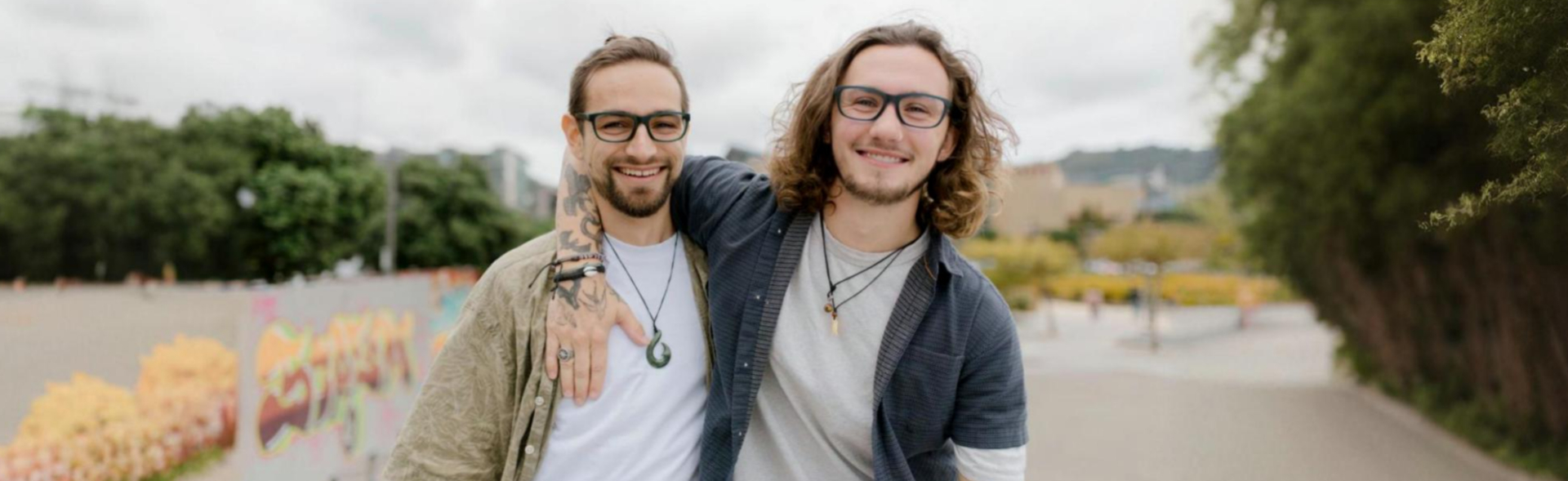 Two men wearing fashionable men's glasses, standing outdoors in a casual setting with graffiti on the walls, greenery in the background, and cloudy skies above. Both are smiling and have their arms around each other.