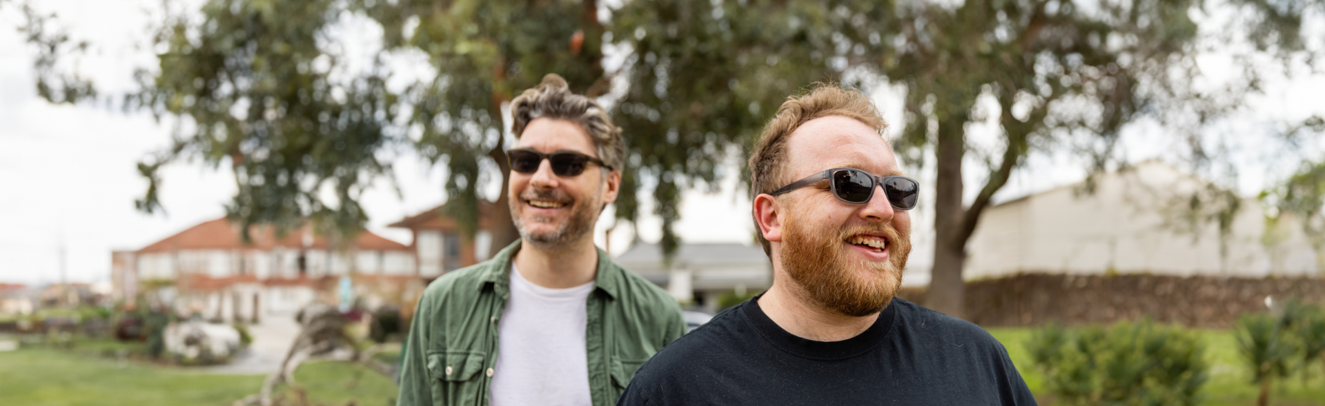 Two men wearing stylish men's sunglasses in Australia, one in a green button-up shirt over a white T-shirt and the other in a black T-shirt, smile outdoors with trees and suburban buildings in the background.