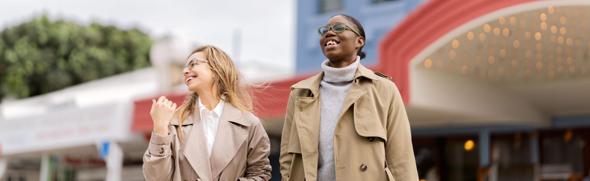 Two women wearing stylish women's glasses from Dresden Vision, walking outdoors in neutral trench coats, smiling confidently against a colourful urban backdrop.