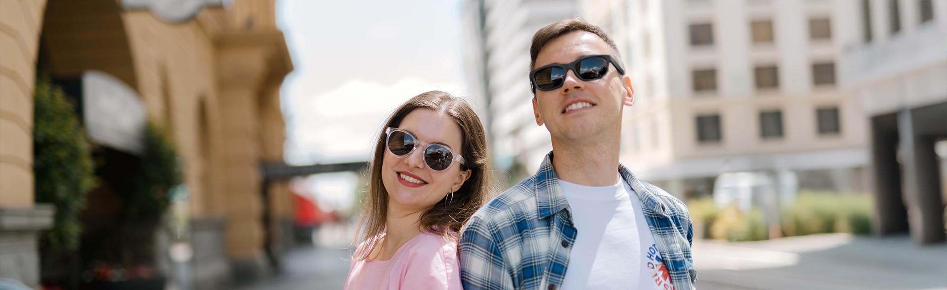 A couple on a city street in the United States, smiling as they wear contrasting sunglasses. The woman has round clear frames, and the man sports rectangular black frames, showcasing Dresden Vision United States' eyewear collection.