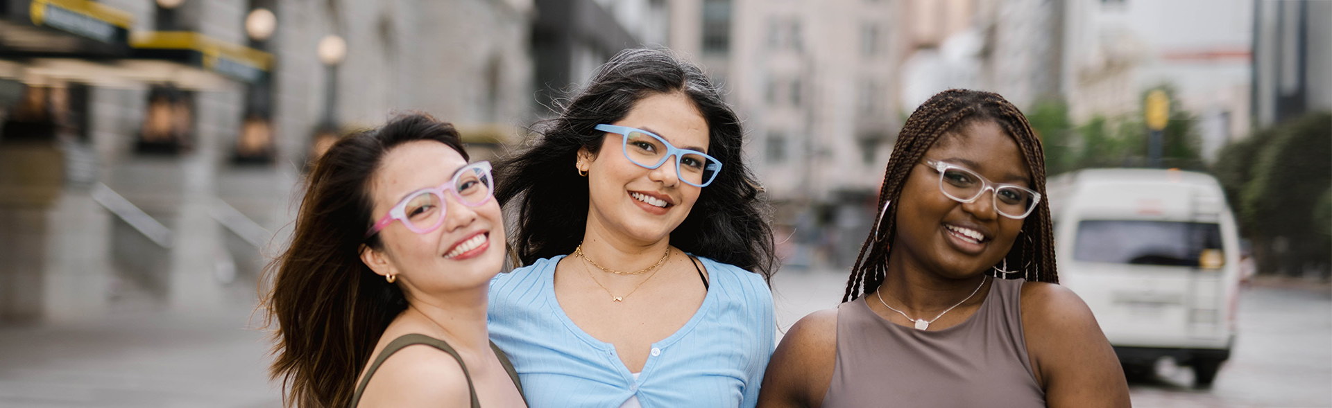 Three friends smiling on a city street, each wearing stylish eyeglasses - one with round pink frames, one with rectangular blue frames, and one with rectangular clear frames, representing Dresden Vision Canada.