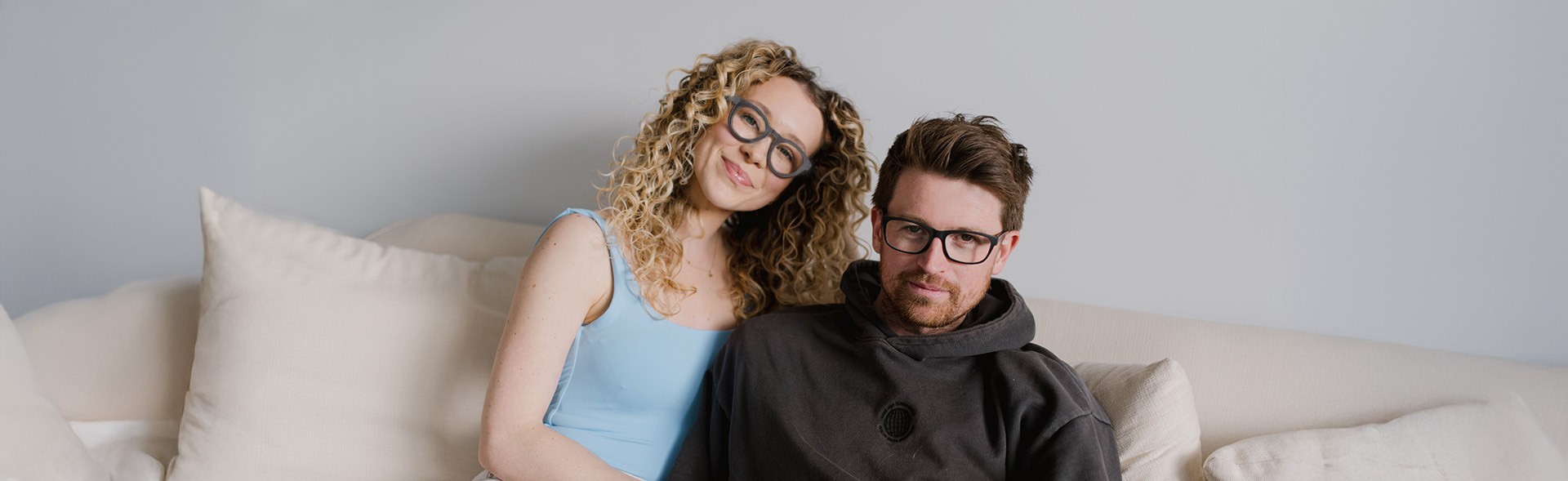 A couple sitting on a white couch, one wearing round grey shaped eyeglasses and the other has black rectangular shaped eyeglasses. She has curly blonde hair and a light blue top, while he is in a dark hoodie, with stylish frames from Dresden Vision United States.